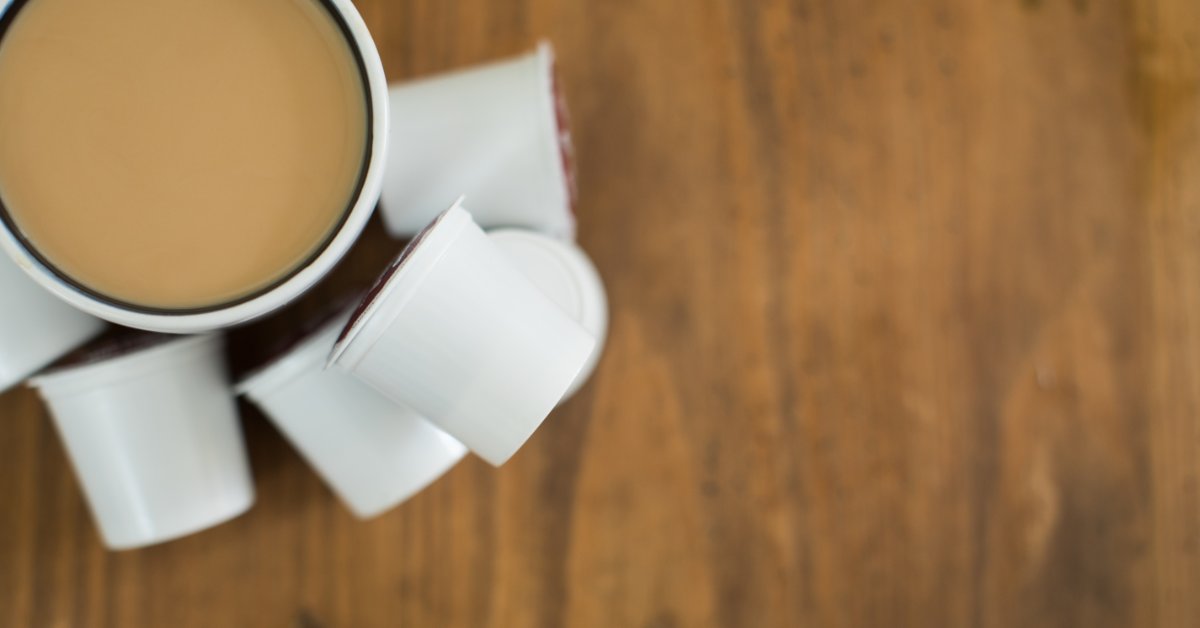 An aerial view of a cup of coffee with cream in it on a wooden tabletop. There are a handful of single-serve pods next to it.