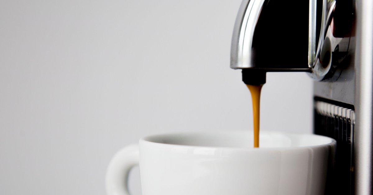A close-up of a single-serve coffee machine brewing hot coffee into a white coffee mug. The spout is black.