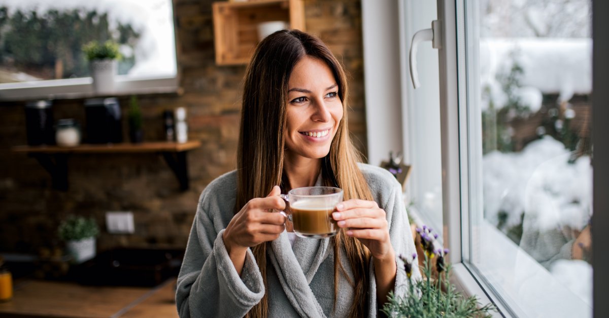 How Coffee Sample Packs Help You Find the Best Iced Brew A brunette woman stands at the kitchen window in a gray robe as she enjoys a cup of hot coffee on a cold day.