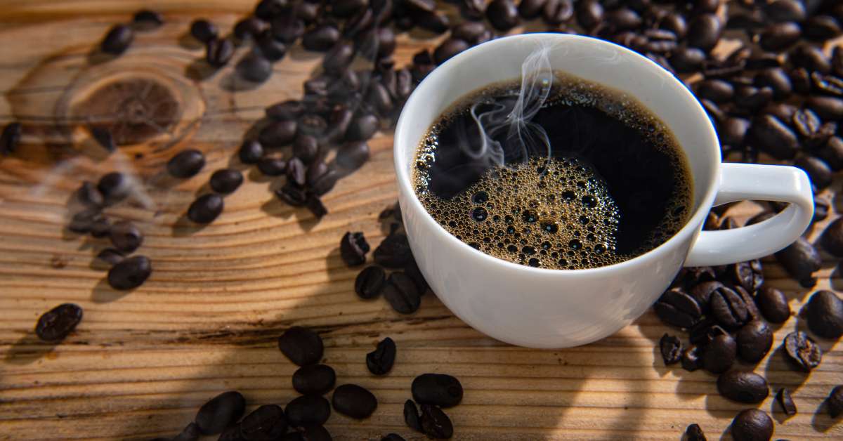 A steaming cup of black coffee sitting on a wooden table and surrounded by loose dark roast coffee beans.