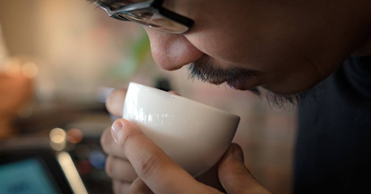 A young bearded man with glasses holds a small white coffee cup up to his nose as if he is smelling it.