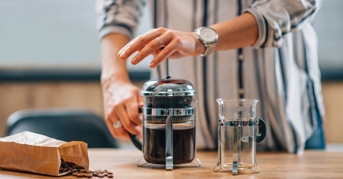 A person wearing a striped shirt and a silver watch uses a French press coffee maker on a wooden table.