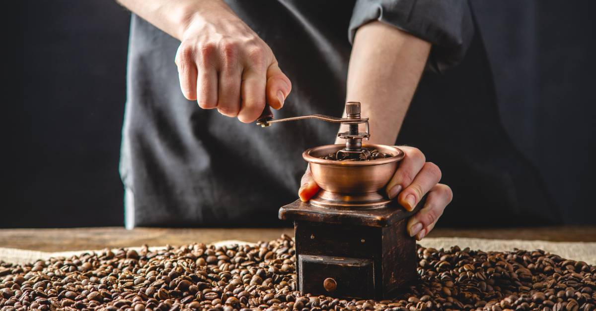 Close-up shot of a person's hands as they use a hand grinder to grind whole coffee beans on a table full of beans.