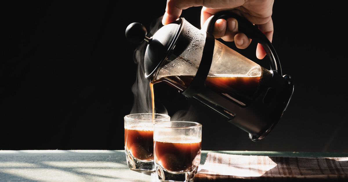 A person holding a French press coffee pot pours fresh coffee into two small glass cups sitting on a table.