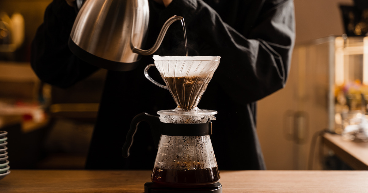 A person wearing a black shirt uses a metal tea kettle to pour water over coffee in a pour over coffee machine.