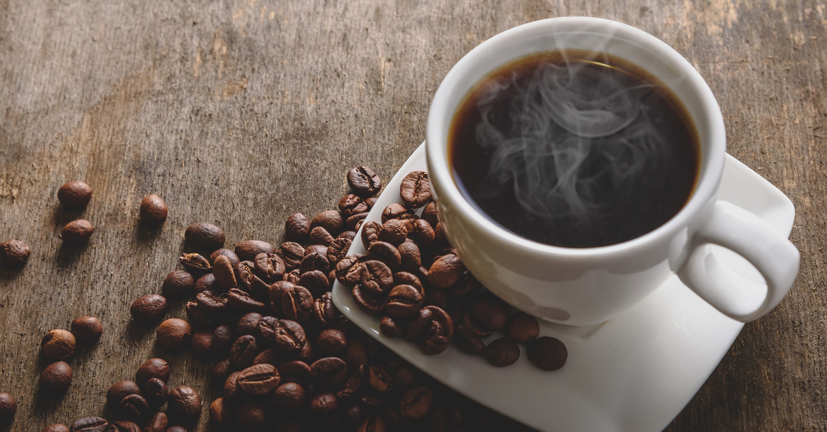 A cup of black coffee in a white mug on top of a white saucer emits steam while surrounded by coffee beans.