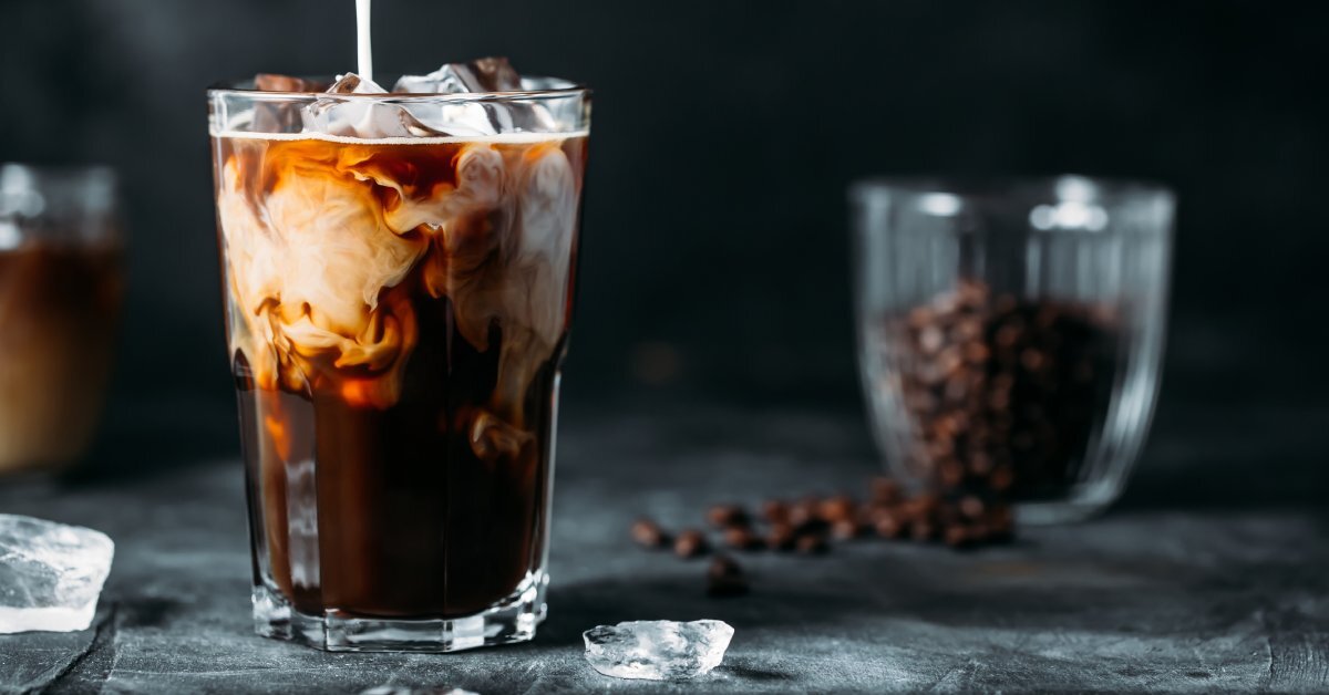 A stream of white milk pours into a small glass filled with ice cubes and black coffee against a black background.