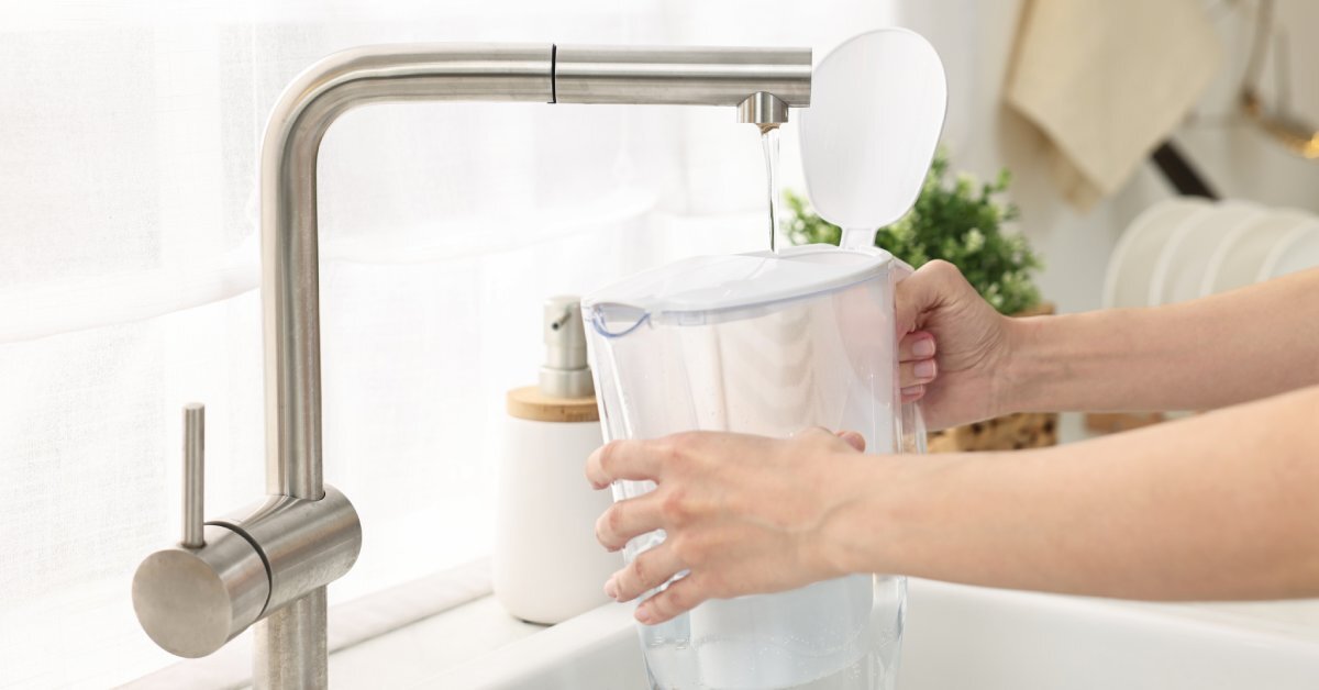 A close-up showing someone holding a plastic water filter jug under a running water faucet to fill it up.