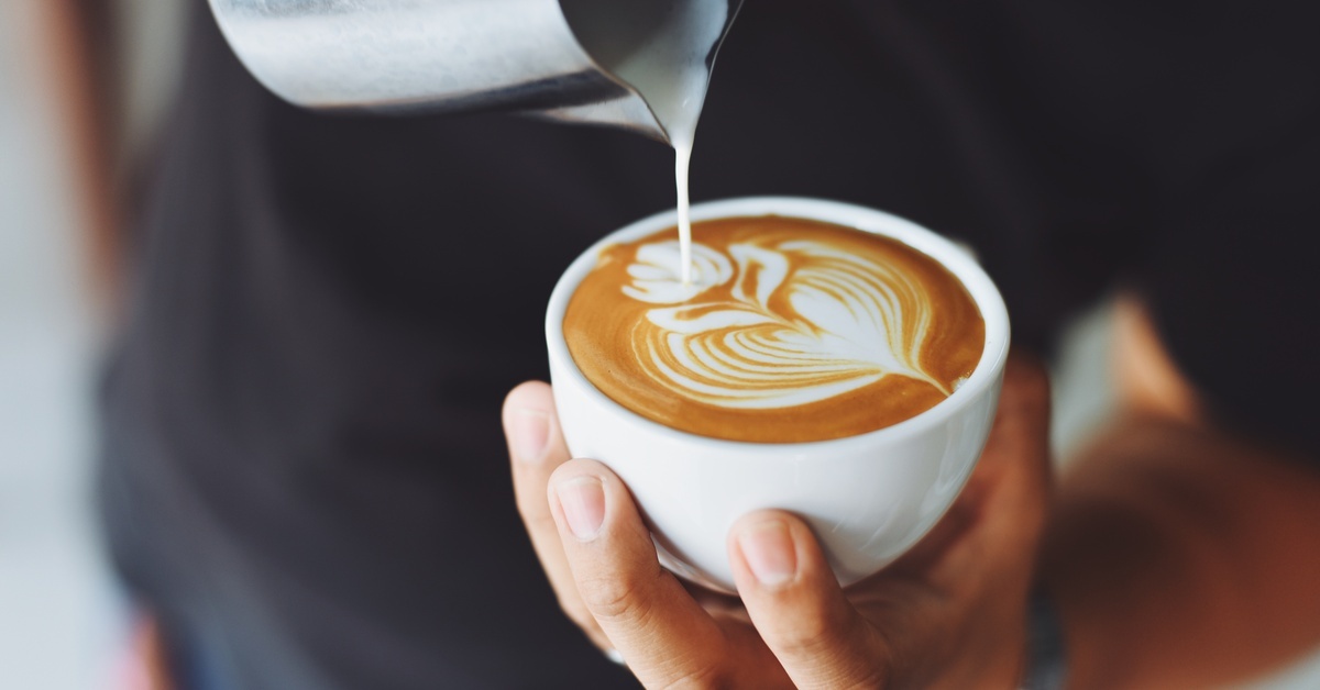 A coffee drinker making their own latte. They're holding the mug in their left hand and pouring frothed milk with the other.