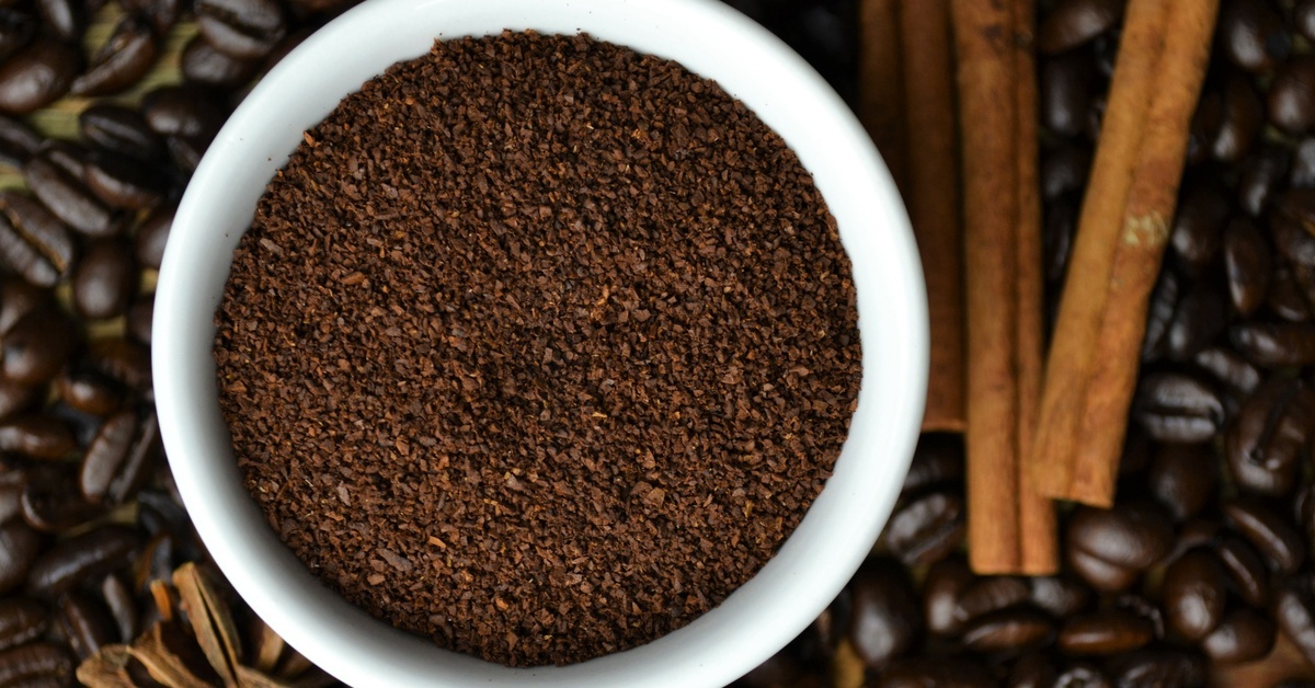 A mug of coffee grounds sitting on a pile of coffee beans. There are two cinnamon sticks laying next to the mug.