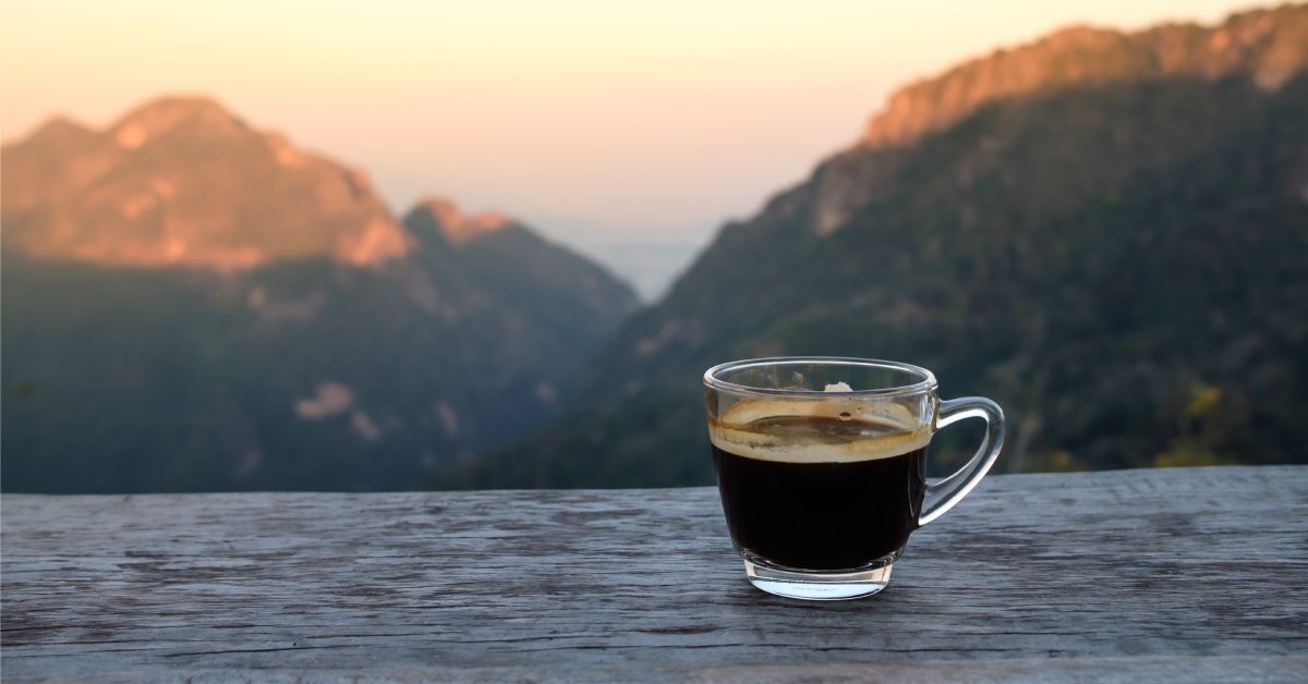 An espresso in a clear class mug sitting on a wooden balcony ledge that overlooks a beautiful mountain range.