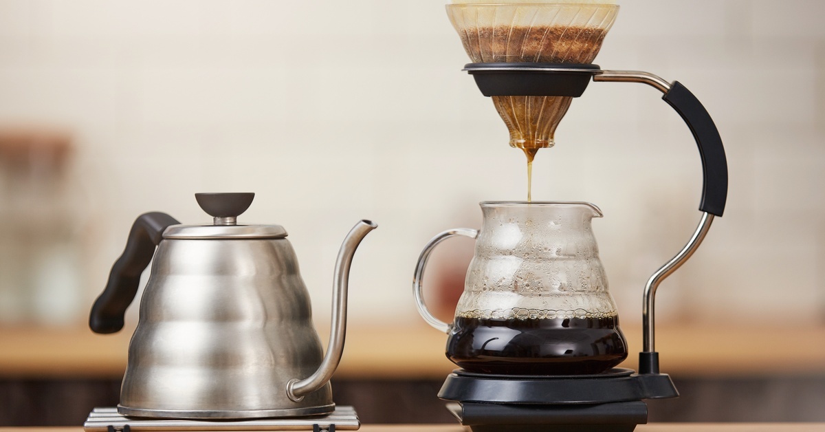 A stainless steel tea kettle with a coffee brewing system to the right. There's a filter with grounds brewing coffee.