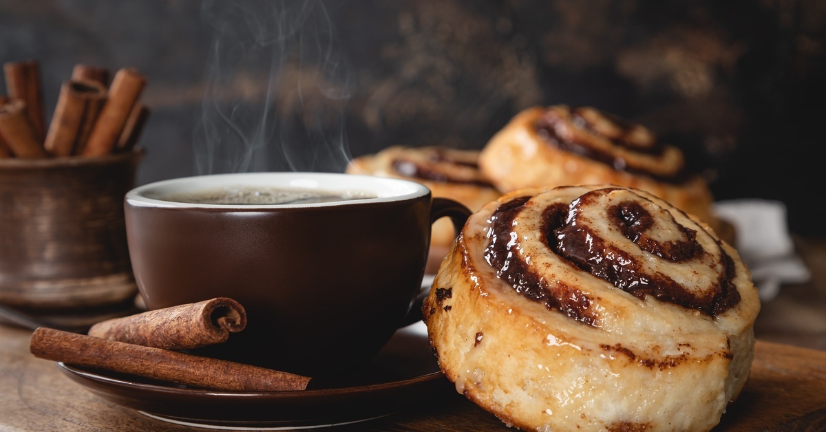 A cinnamon roll sits on the edge of a plate next to a dark brown coffee mug. There are two cinnamon sticks by the mug.