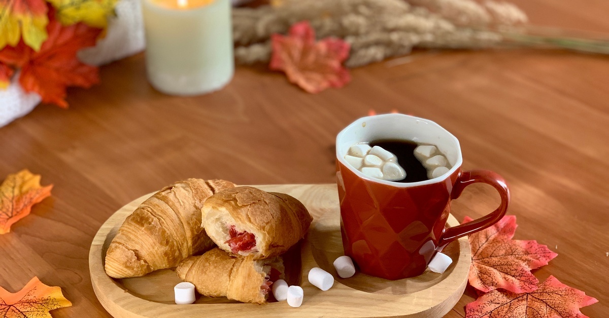 A table decorated with fall leaves and a wooden tray with a cup of coffee and flakey rolls. The rolls have red filling.