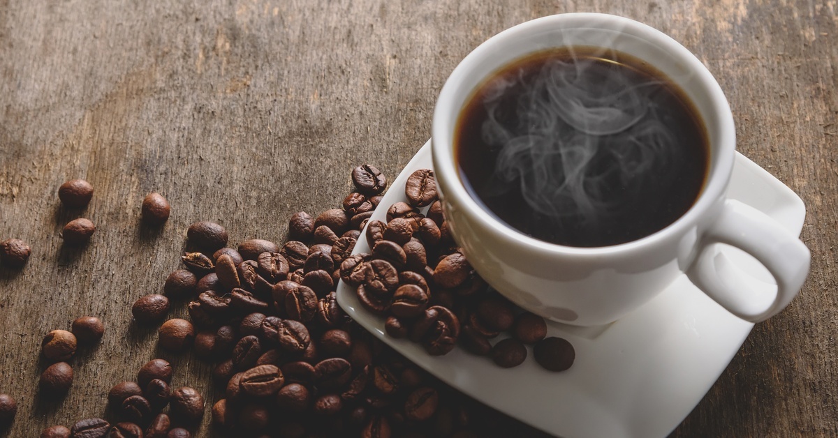 What You Need To Know About Single-Origin Coffee Beans A cup of black coffee sits on a saucer on the table next to a pile of loose coffee beans. The table is wood.