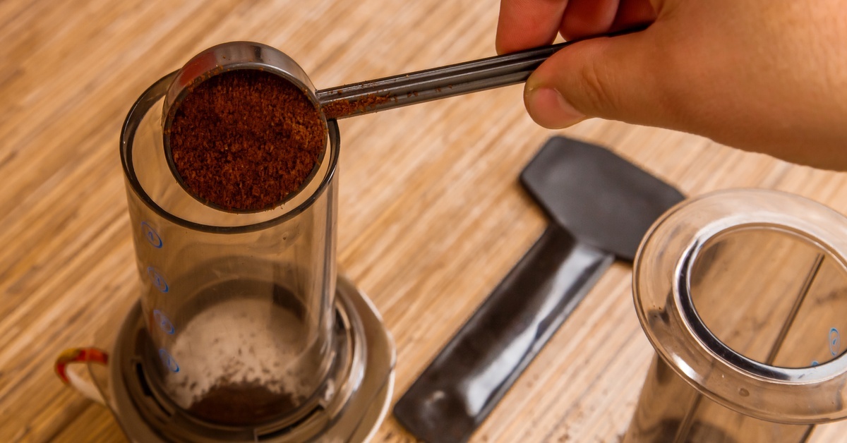 A close-up of a hand using a coffee scoop to drop finely ground coffee into an AeroPress brew system. A close-up of a hand using a coffee scoop to drop finely ground coffee into an AeroPress brew system.