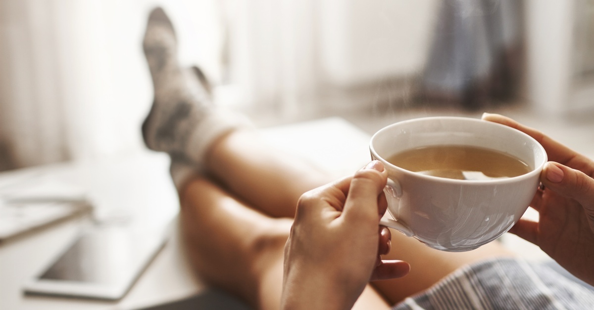 A person at home with their feet on the coffee table. They're holding a white coffee mug with both hands. A person at home with their feet on the coffee table. They're holding a white coffee mug with both hands.