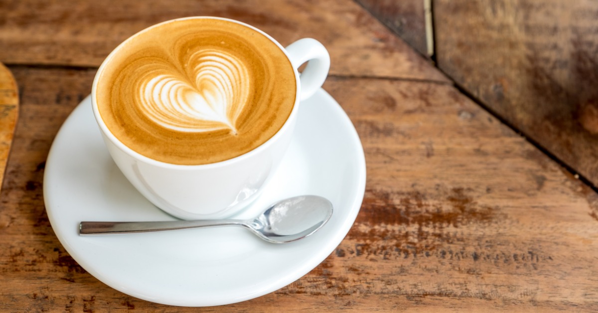 A cup of coffee in a white mug sits on a white saucer. The coffee is frothy with a white foam heart in the top.