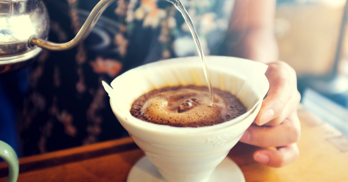 A person at the counter using a tea kettle to pour hot water over a basket of coffee grounds in a pour-over system.