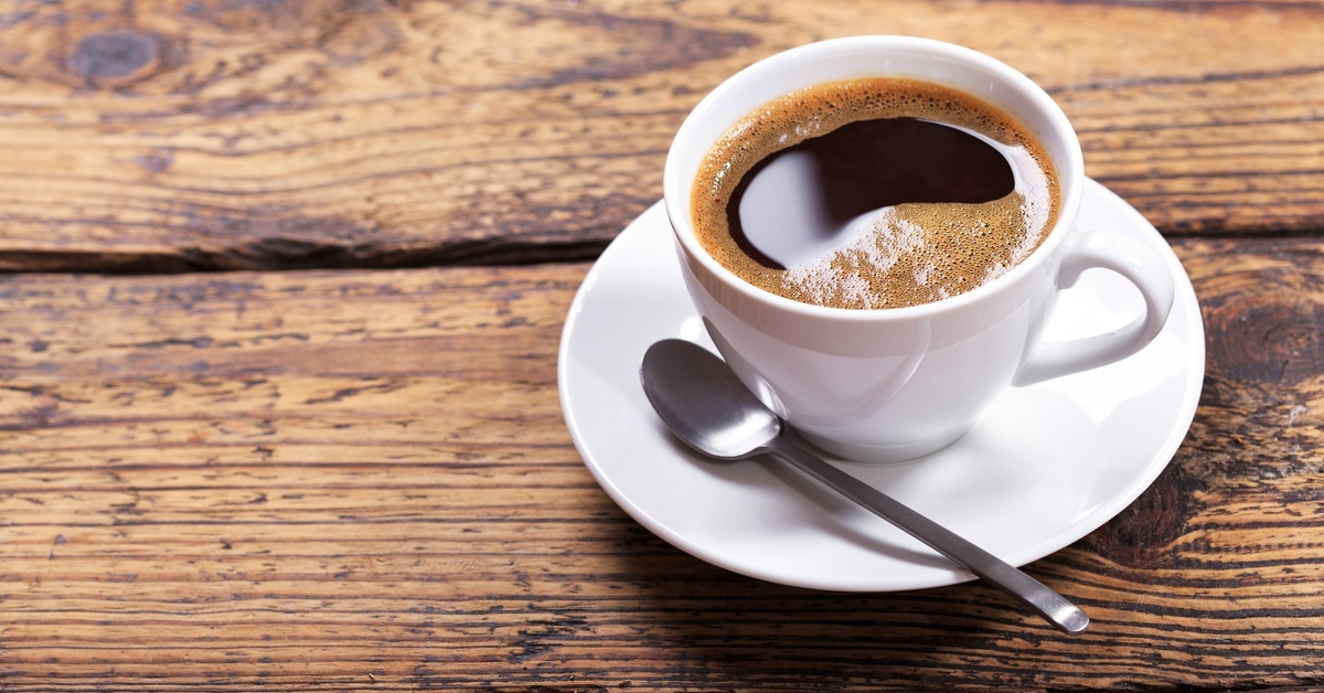 A cup of black coffee with froth in a white mug and saucer with a silver spoon sits on a wooden tabletop.
