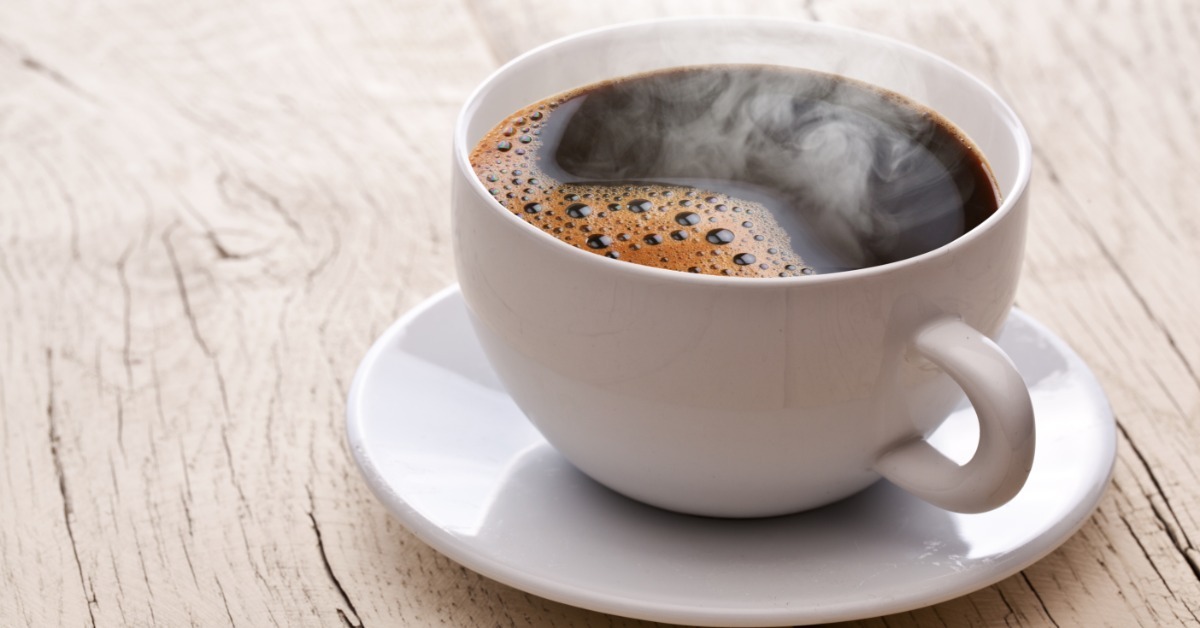 A white coffee mug sits on a matching saucer with black coffee sitting in the mug. There is steam coming off the coffee.