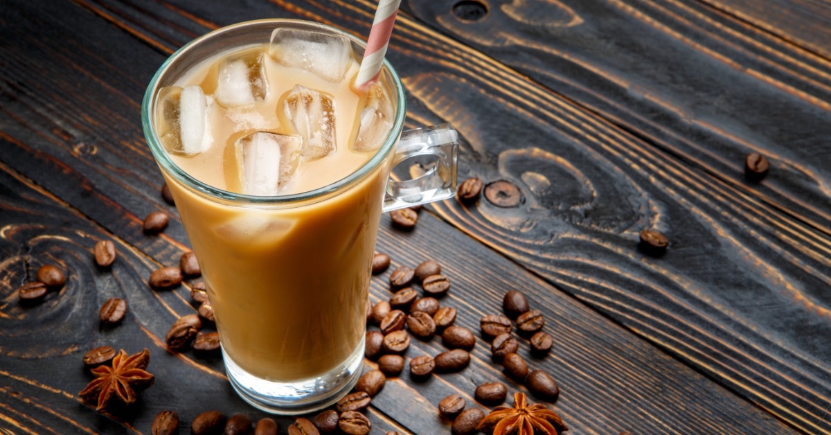 An iced coffee sits on a table with a straw in the glass. There are coffee beans scattered around the glass.