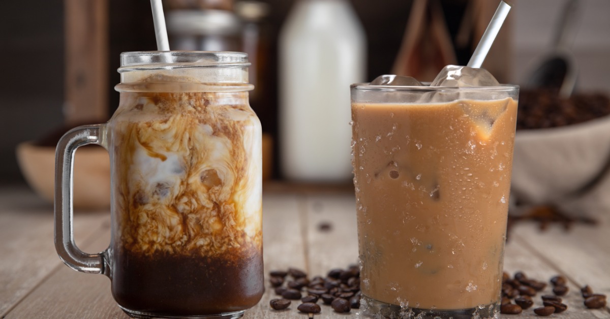 Two glasses of iced coffee sit on the countertop. They each have milk swirled throughout and a straw in the glass.