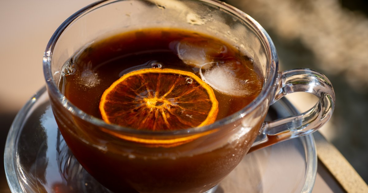 A close-up of a clear glass coffee mug with black coffee and a slice of lemon floating on top. There are a few ice cubes.