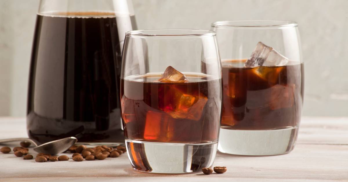 Two glasses of cold brew with ice sit next to a glass carafe with cold brew in it. There are beans on the table.