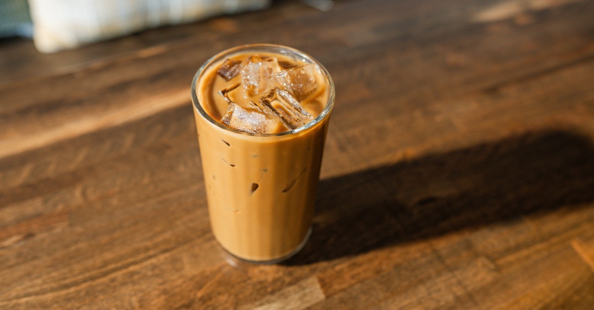 An iced coffee sits isolated on a wooden tabletop surface. The drink sits in a glass and is a milky brown color. An iced coffee sits isolated on a wooden tabletop surface. The drink sits in a glass and is a milky brown color.